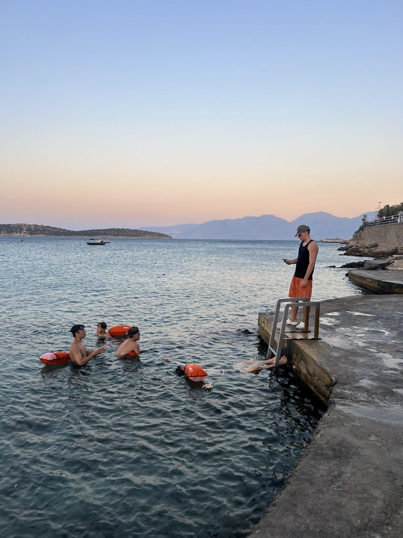 Campers swimming in the sea with orange buoys while a coach watches from the pier