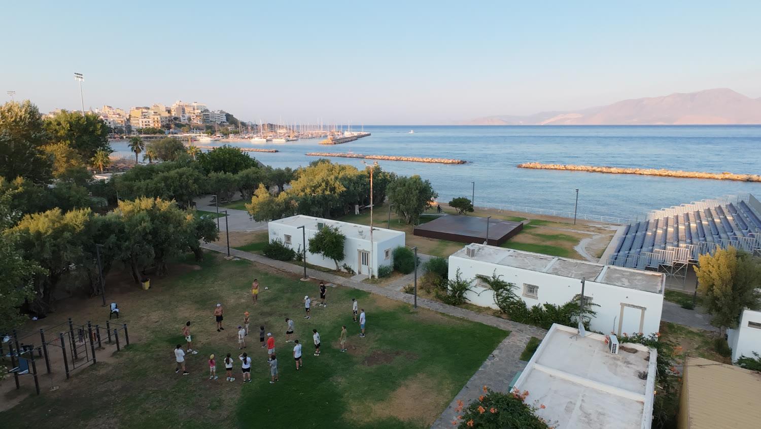 Drone view of the grounds by the sea: white buildings among trees, the group warming up on the lawn