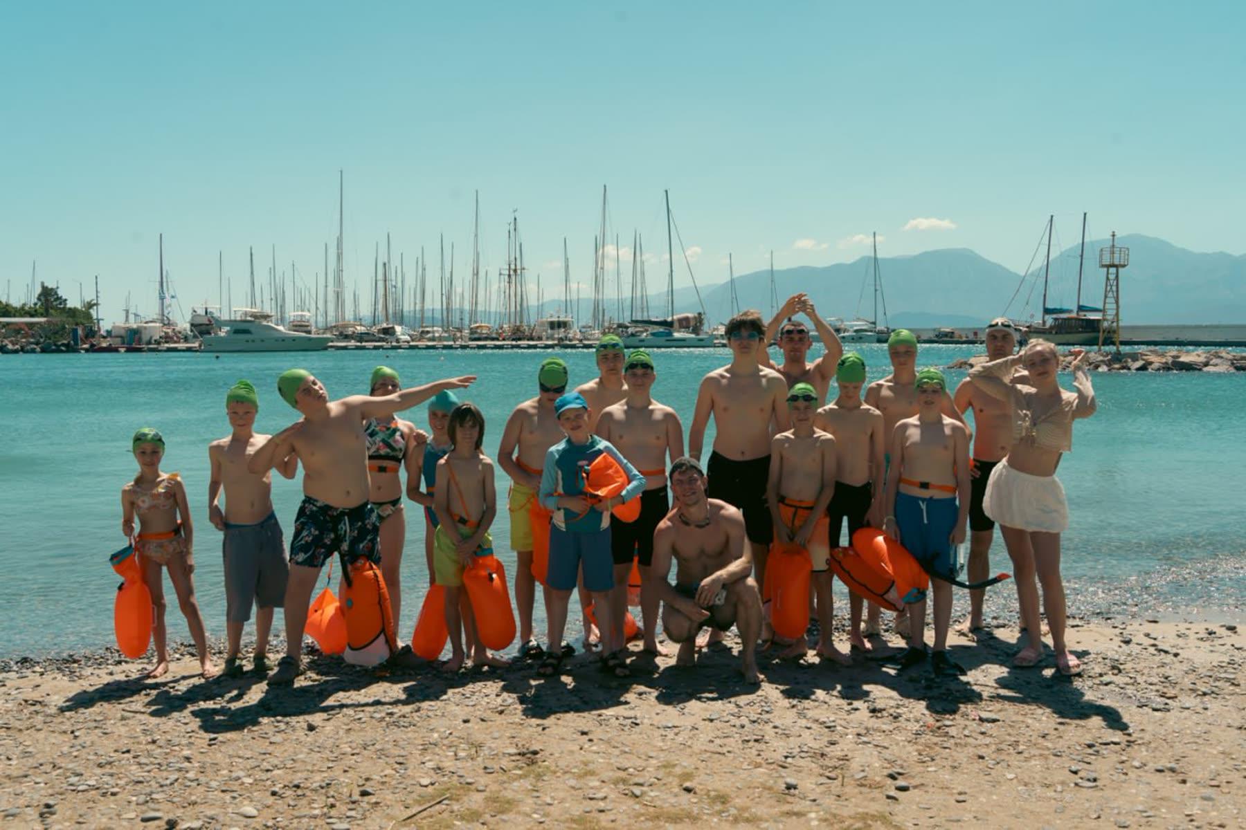 The camp group lined up on a beach with orange buoys, a harbour behind them